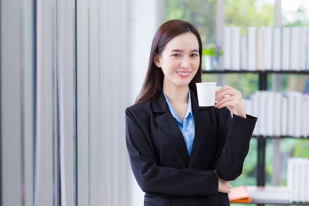 Asian business female who wears blue shirt and black suit holds coffee cup in her hand and smile