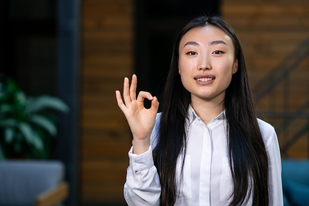 Asian business woman smiling and showing OK sign standing in the hallway in the office