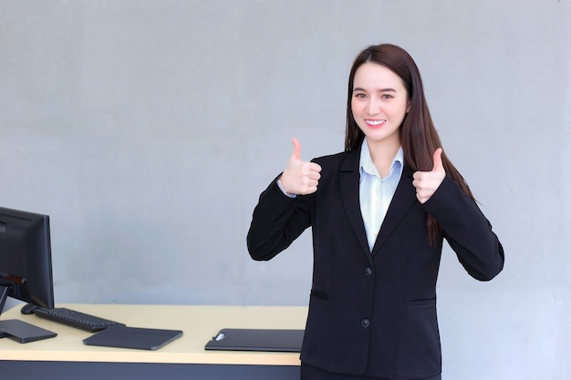 Asian business woman who wears black suit  smiles and shows hand thump up as good sign in the office