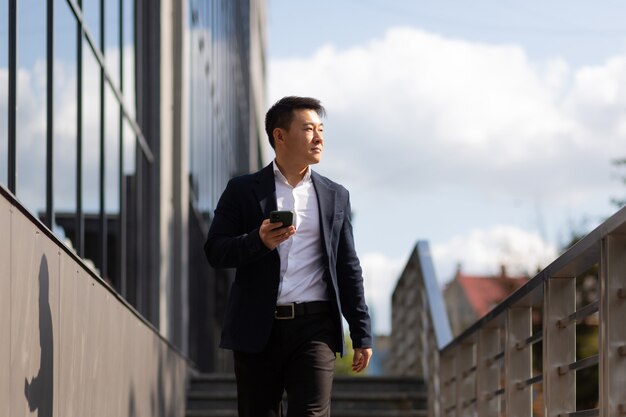 Photo asian businessman boss looks at mobile phone reads news from phone and writes in messenger outside office