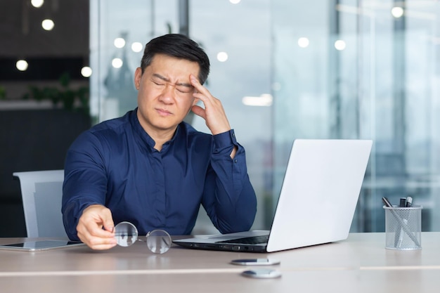 Asian businessman boss sick has severe headache man in glasses sitting at desk using laptop at work