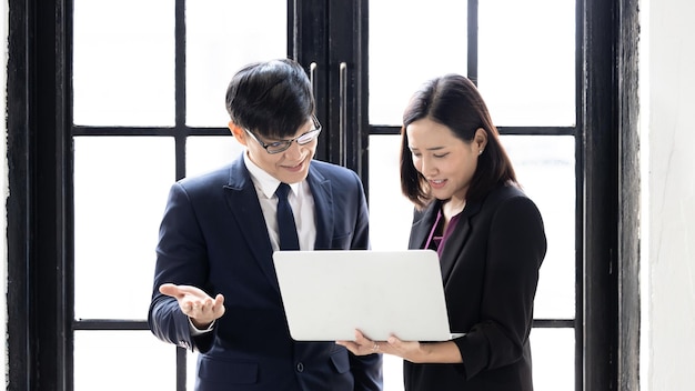 Asian businessman and businesswoman startup using computer laptop together while standing in office building windows Successful young business partners in suits looking at laptop strategic analysis