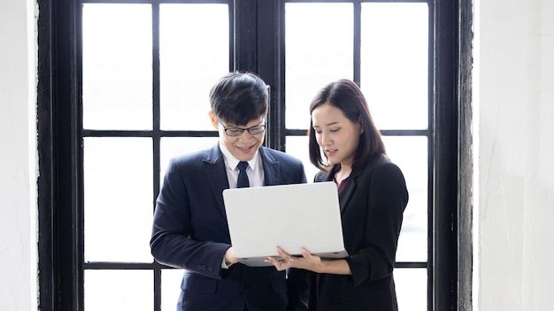 Asian businessman and businesswoman startup using computer laptop together while standing in office building windows Successful young business partners in suits looking at laptop strategic analysis