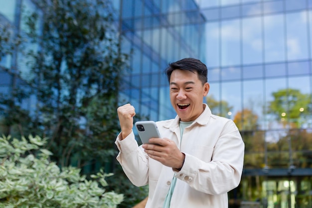 Asian businessman in casual clothes with phone outside office building celebrating victory and