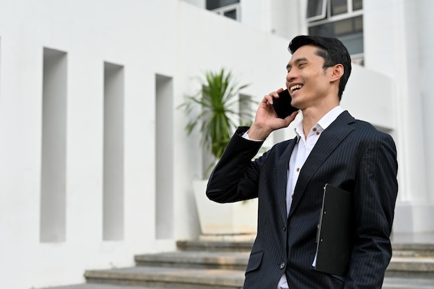 Asian businessman in formal suit standing outside of the building and talking on the phone