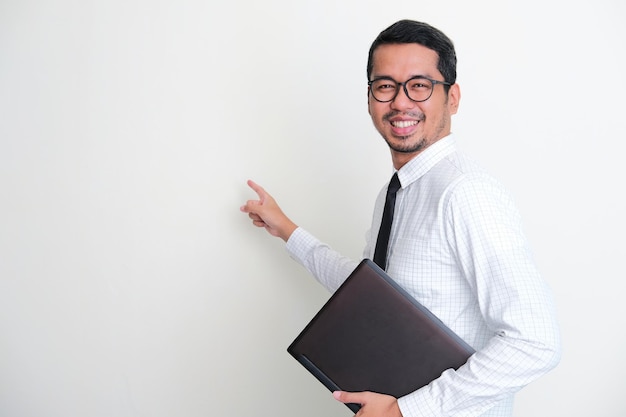 Asian businessman holding a laptop pointing behind with confident expression