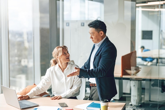 Asian businessman is explaining plans to mature caucasian woman that working on laptop in office