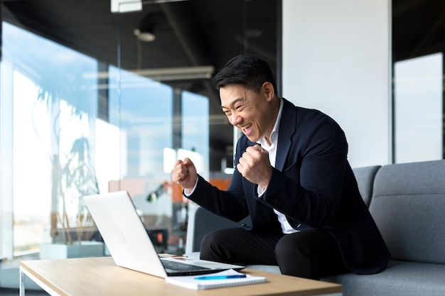 ASIAN Businessman rejoices in success looks at the laptop screen happy and smiles got a great result of his work celebrates victory sitting in the office