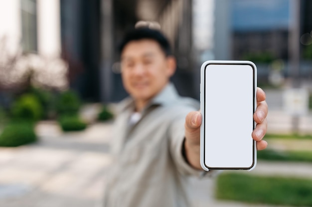 Asian businessman showing smartphone with blank screen to camera posing outdoors near office building mockup