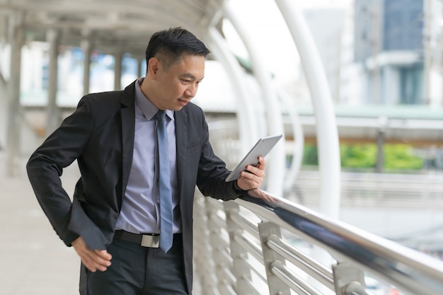 Asian Businessman standing and holding digital tablet with business office buildings