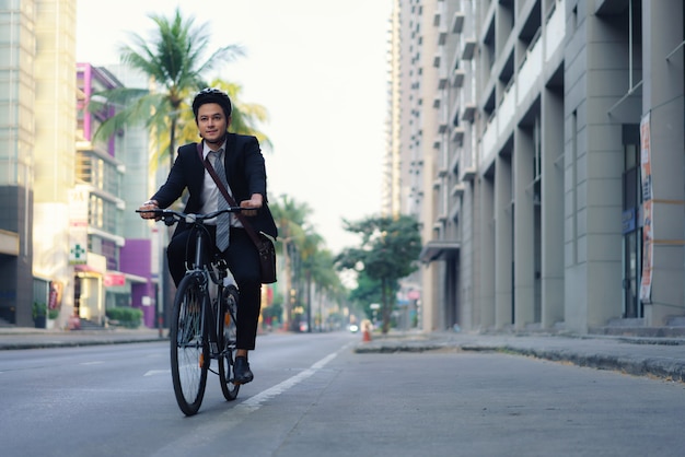 Asian businessman in a suit is riding a bicycle on the city streets for his morning commute to work. Eco Transportation Concept.
