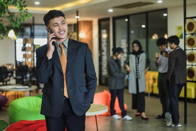 Asian businessman talking on the phone cheerful smiling reports good news, against blurred coworker