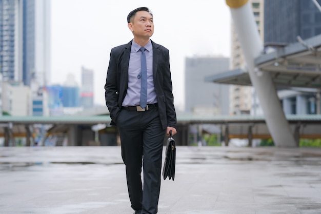 Asian Businessman walking and holding briefcase with business office buildings in the city