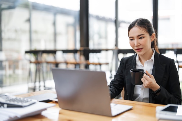 Asian businesswoman in formal suit in office holding mug working on laptop at office