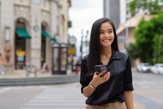 Asian businesswoman outdoors in city street using mobile phone while walking and smiling