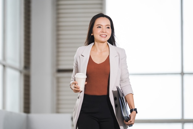 Asian businesswoman in a relaxed business suit holds take away coffee cup stands in company building