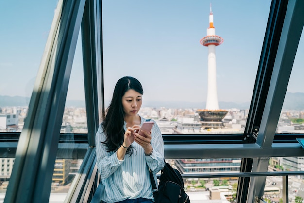 asian businesswoman standing indoor in train station with the kyoto tower in the background. young office lady checking the timetable of the transport on cellphone. beautiful worker visiting abroad.