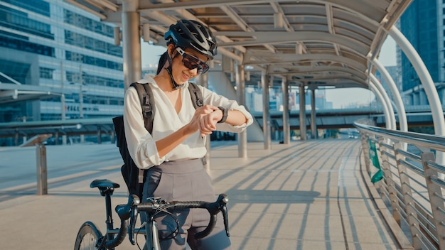 asian businesswoman with backpack bicycle looking smartwatch in city street