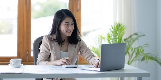 Asian businesswoman working in financial accounting analyzes a chart using a laptop calculator with documents and holding a pen sitting on a chair at an office desk