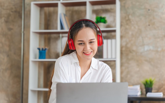 Asian businesswoman working on laptop computer and wearing stereo headphones to listen to music work remotely live streaming relaxed concept