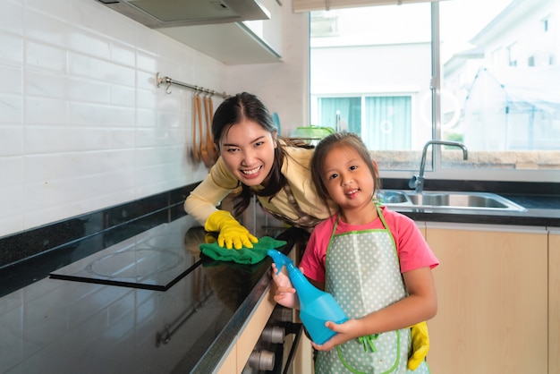 Asian Cheerful mother and daughter cleaning table surface with rag and spray bottle together at kitchen counter in house.