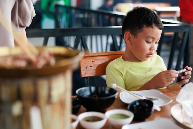 Asian child boy playing online game on smartphone at restaurant while waiting mom grill food