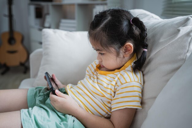 Asian child girl looking using and touch mobile phone screen on couch sofa Baby smiling funny time to use mobile phone Too much screen time Cute girl watching videos while tv Internet addiction