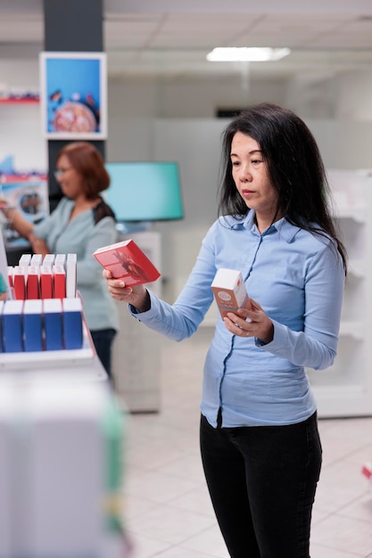 Asian client wanting to buy medicaments in health care pharmacy, looking at medicinal products and pharmaceutical supplies. Woman in medical drugstore with pills boxes on shelves.