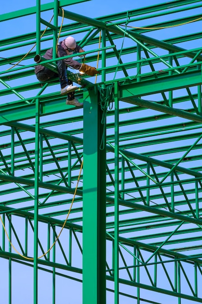 Asian construction worker is welding metal on green roof warehouse structure against blue sky