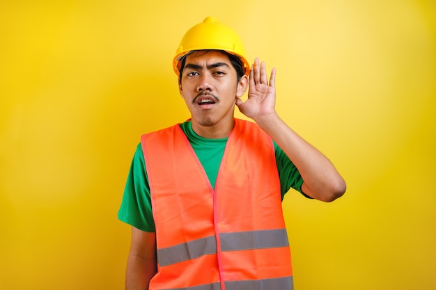 Asian constructor man wearing orange vest and safety helmet over yellow background with hand over ear listening an hearing to rumor or gossip. Deafness concept.