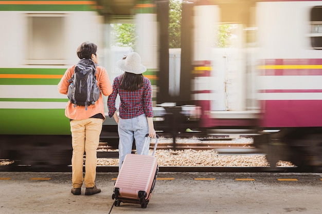 Asian couple tourists with backpacks standing on platform of railway in the train station with train movement is background.