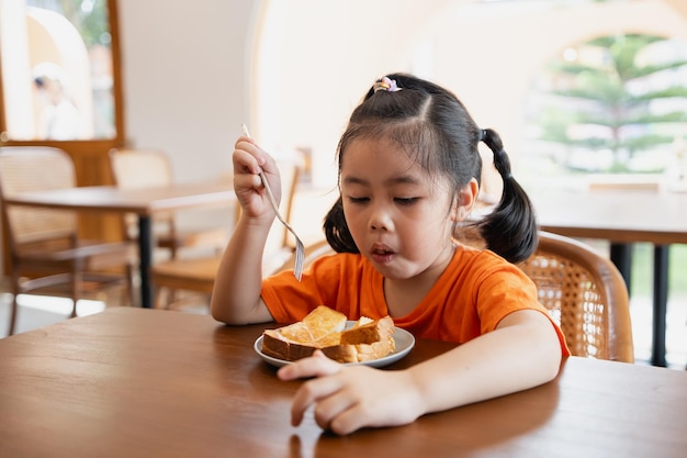 Asian cute baby child girl sits at a table savoring a delicious breakfast With a smile on her face she enjoys eating toast and experiencing the charm of the quaint restaurant In a cozy cafe