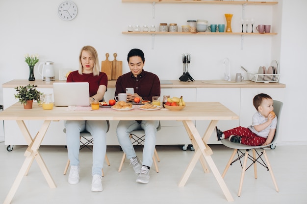 Asian dad and european mom work with their gadgets, and son has breakfast alone. Modern technological problems.