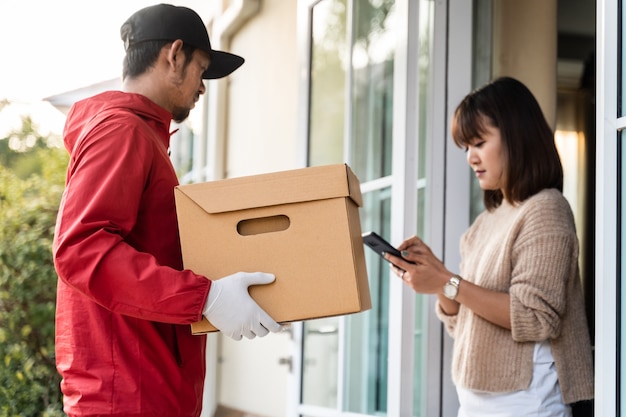 An Asian deliver man in red uniform handing parcel to a female costumer in front of the house. A postman and express delivery service deliver parcel during covid19 pandemic.