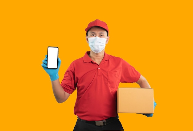 Asian delivery man worker showing mobile phone on white screen wearing surgical mask medical gloves in red uniform isolated on yellow background using mobile checking stock and hold parcel boxes