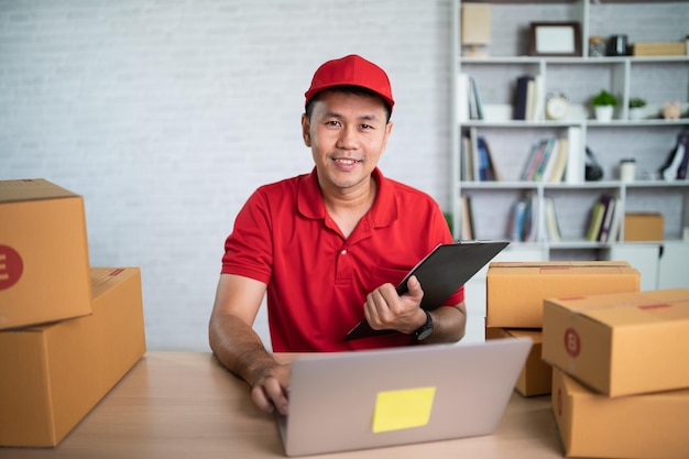 Asian delivery man worker smiling in red uniform work at home checking list parcel boxes for sending or conveying parcels by mail Delivery transport concept