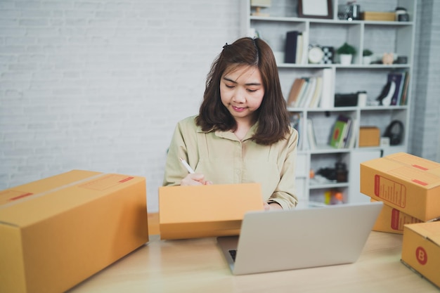 Asian delivery woman freelance working at home checking list parcel boxes for sending or conveying parcels by mail