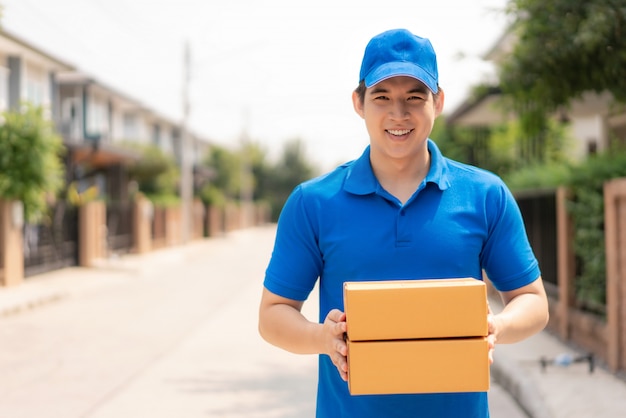 Asian delivery young man in blue uniform smile and holding pile of cardboard boxes in front house village with copy space.