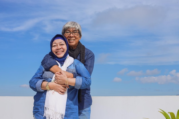 Asian elderly couple in casual style is smiling old man hug his wife over blue sky background