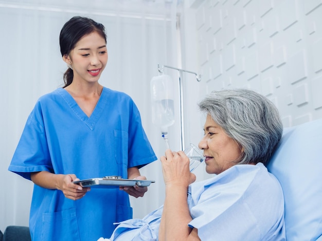 Asian elderly patient taking medicine hold drinking water glass and pill from young nurse in blue scrub suit in hospital room daily medicine or vitamin supplements senior healthcare and medical