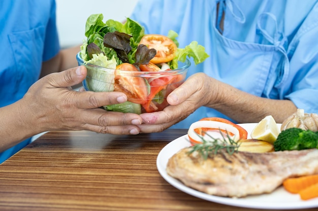 Asian elderly woman patient eating Salmon steak breakfast with vegetable healthy food while sitting and hungry on bed in hospital