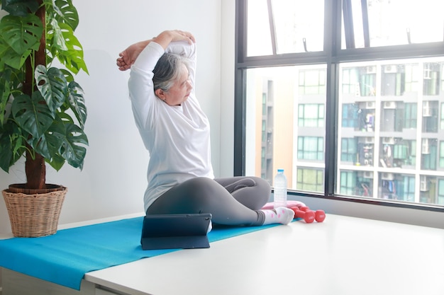 Asian elderly women exercise at home Do arm stretches according to the trainer on the tablet. Via online video. Health care concept for the elderly