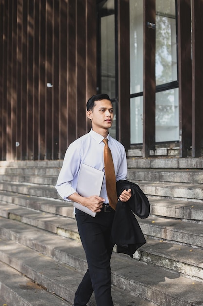 Asian employee walking outdoors outside the building with laptop and suit on his hands.