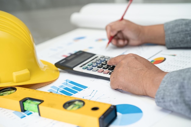 Asian engineer with plan project blueprint graph and yellow helmet and engineering construction tools