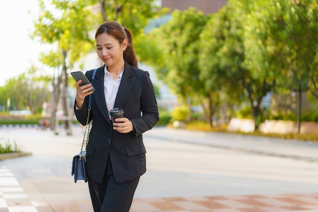 Asian executive working woman holding coffee cup and using a mobile phone in the street with office buildings in the background in Bangkok, Thailand.
