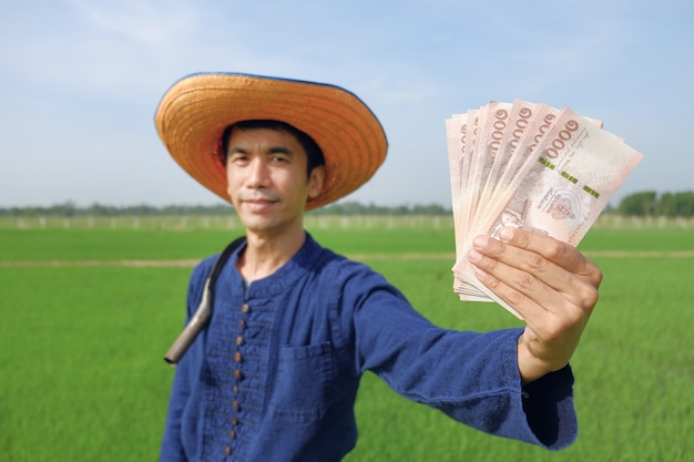 Asian farmer man holding Thai banknote money at green rice farm. Image focus at banknote and blur face.