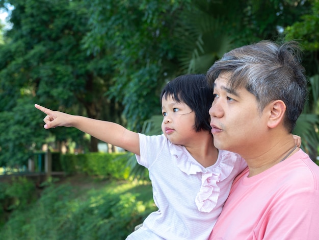 An Asian father is carrying a lovely daughter to her chest. The daughter is pointing out to see what he sees. 