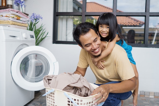 Asian father playing piggy back ride together with daughter while doing laundry using washing machine