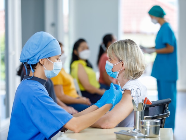 Asian female doctor wears face mask and blue hospital uniform injecting vaccine to Caucasian senior woman at ward working desk with equipment while other patients wait in queue in blurred background.