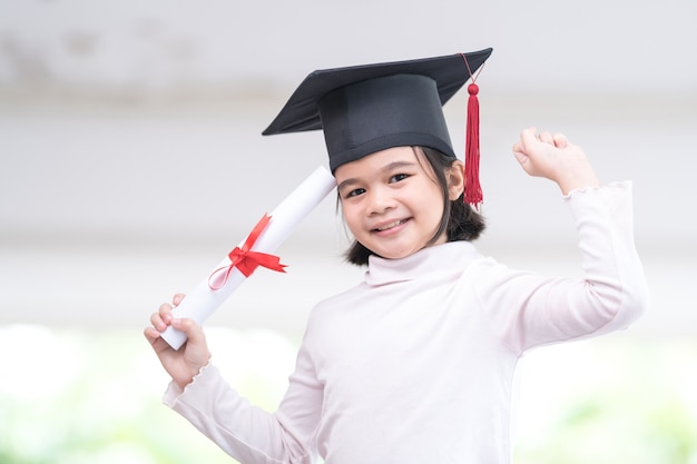 Asian female school kid graduate in a graduation cap holds a rolled certificate paper